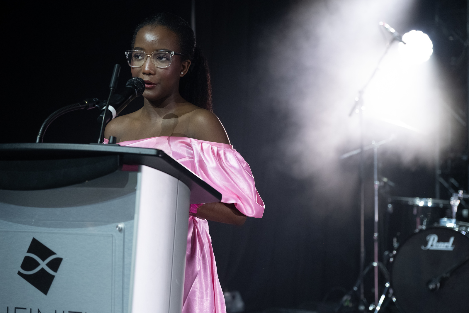 A young woman wearing a pink dress stands at a podium and speaks into a microphone.