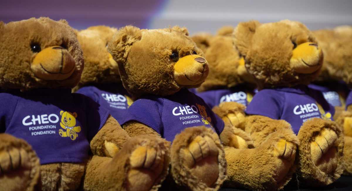 A table of beige teddy bears wearing purple t-shirts with a CHEO logo.