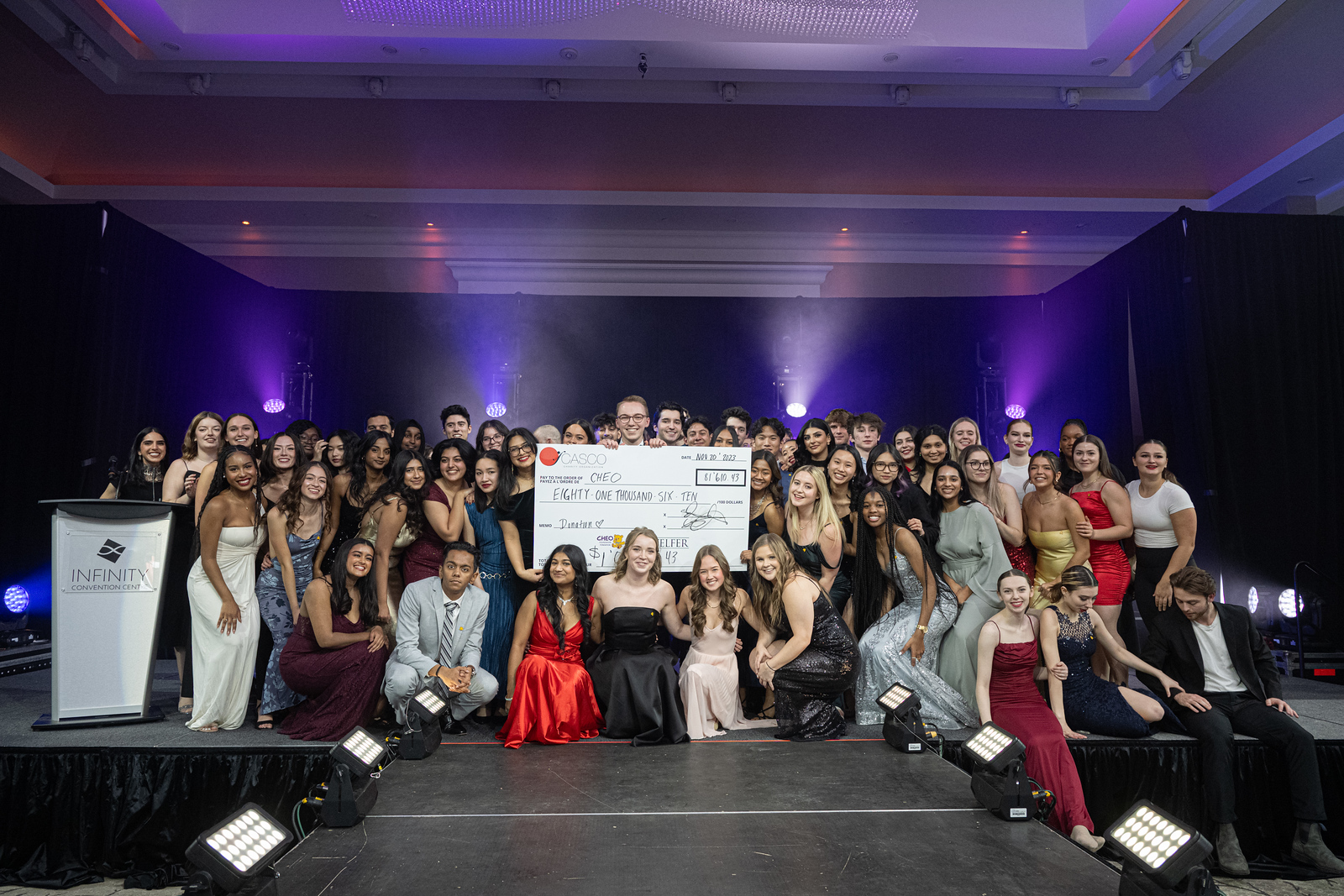 A large group of young adults in formal wear pose for a photo on a stage with a cheque.