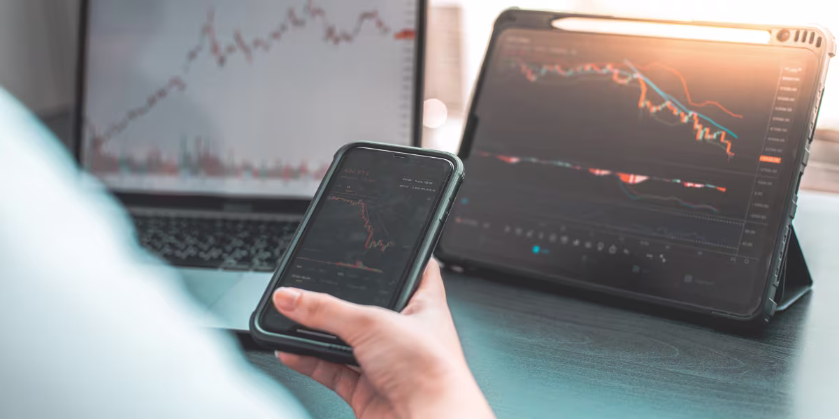 The image shows a person holding a smartphone displaying a stock market chart, while a tablet and a laptop in the background also show financial graphs.