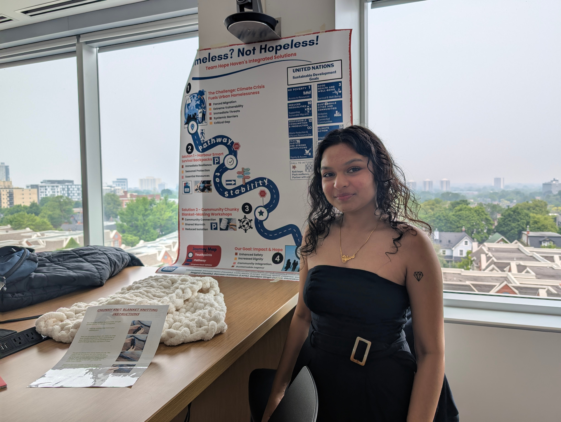 Student standing in front of her homelessness project poster, with a chunky knit blanket prototype on the table.