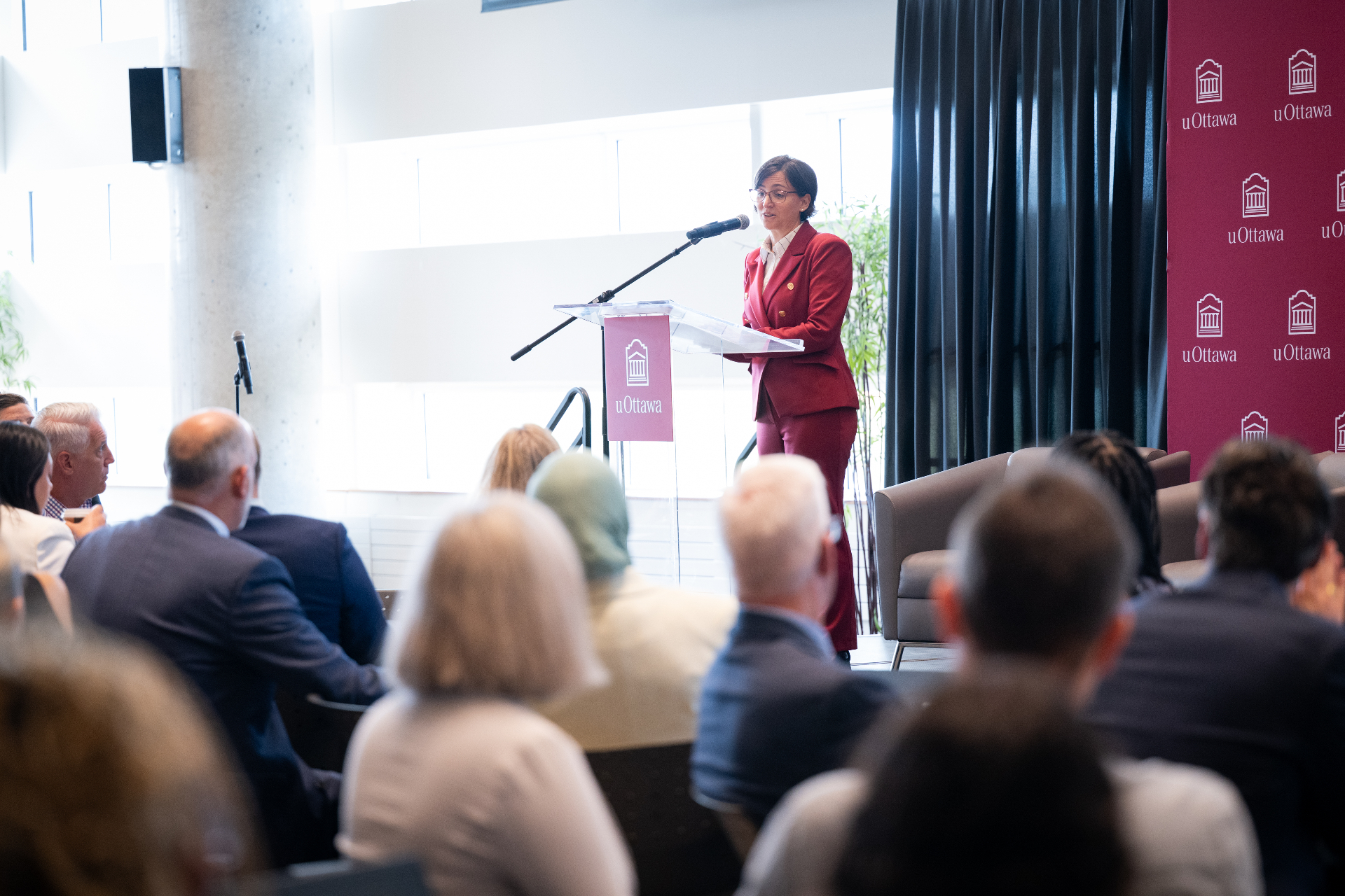 Une femme vêtue d’un tailleur rouge parle à un podium muni d’un microphone, s’adressant à un auditoire. Le podium affiche le logo de l’Université d’Ottawa et l’arrière-plan présente l’image de marque de l’université.