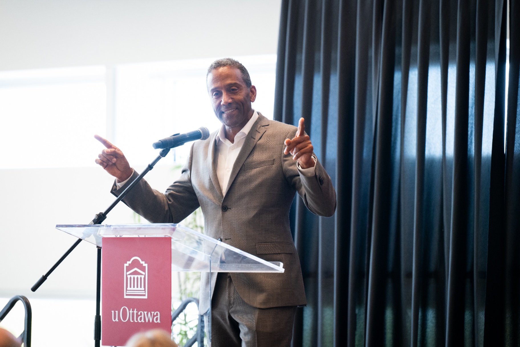Un homme en complet gris parle à un podium muni d’un microphone, en gesticulant avec les deux mains. Le podium arbore le logo de l’Université d’Ottawa avec une toile de fond.