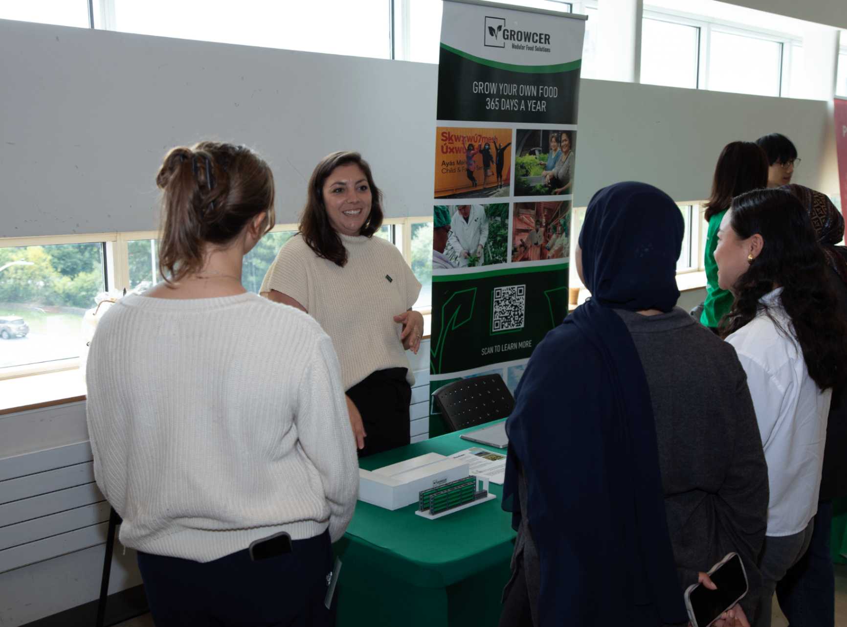 Une femme se tient à un kiosque de Growcer et discute avec un petit groupe d’étudiantes. Le kiosque présente une bannière verte faisant la promotion de la culture alimentaire durable et un code QR pour en savoir plus.