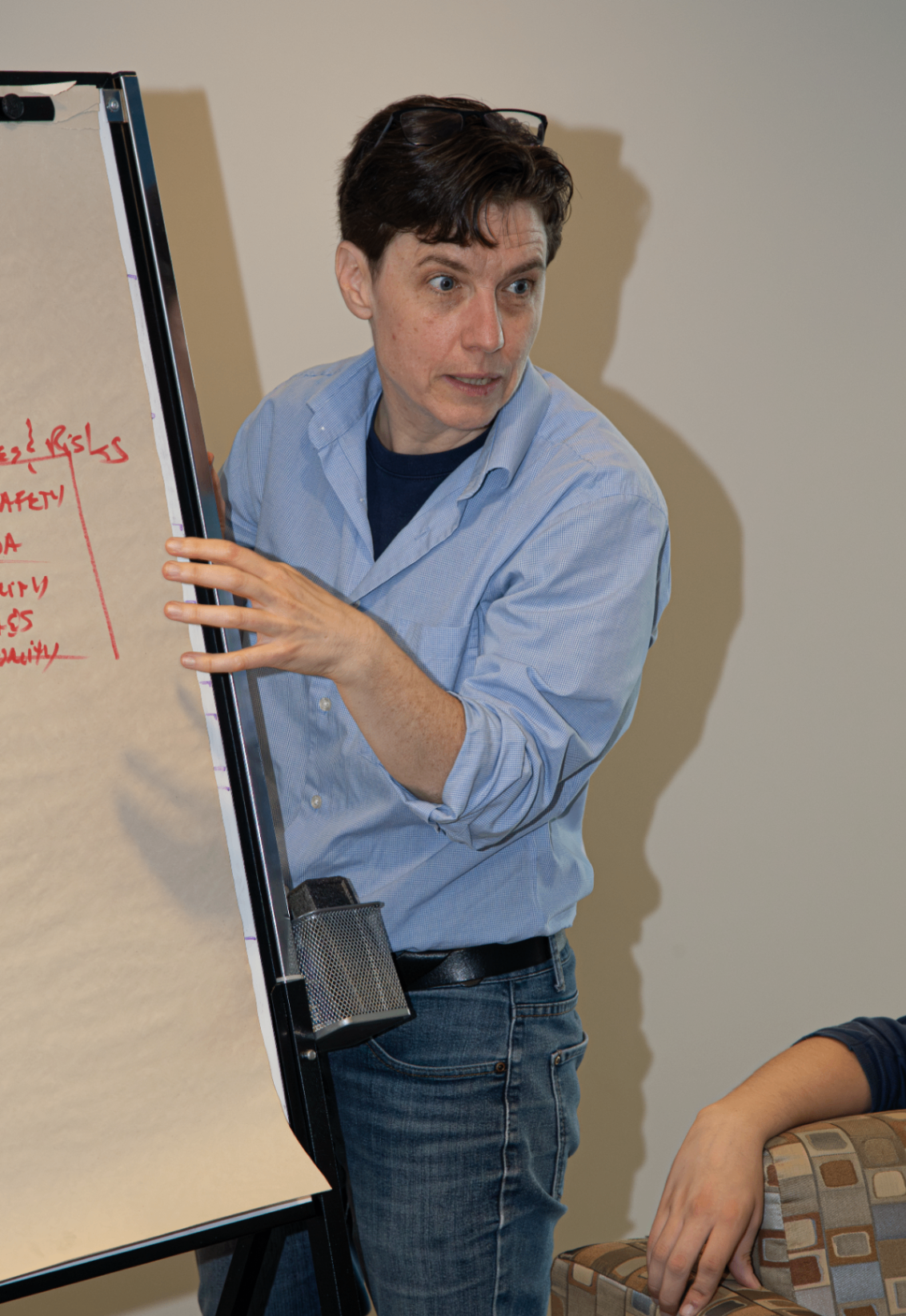 Une personne portant une chemise bleu pâle et un jean présente un tableau à feuilles mobiles couvert de notes manuscrites. Elle semble concentrée et engagée, expliquant un concept à d’autres personnes assises à proximité dans un contexte de discussion.