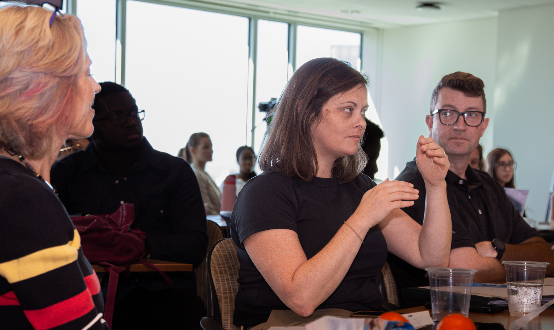 Plusieurs participantes et participants sont assis dans une salle de classe bien éclairée, prenant part à une discussion. Une femme au premier plan parle en faisant des gestes, tandis que les autres écoutent attentivement. L’ambiance reflète un échange interactif lors d’un atelier ou d’une présentation.