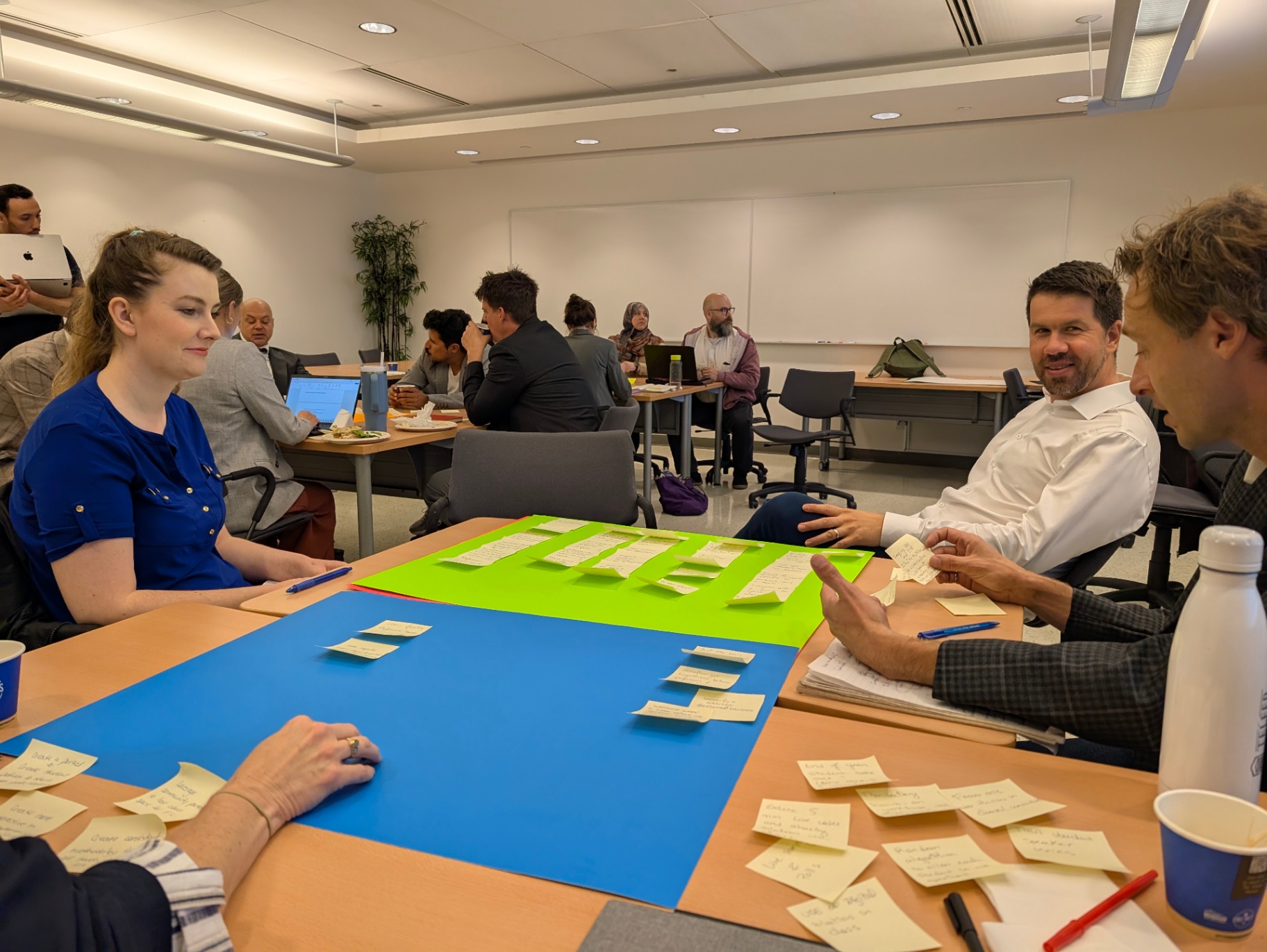 Des participantes et participants sont assis autour d’une table lors d’un atelier, discutant d’idées et organisant des notes autocollantes sur de grandes feuilles bleue et verte. D’autres groupes travaillent en arrière-plan.