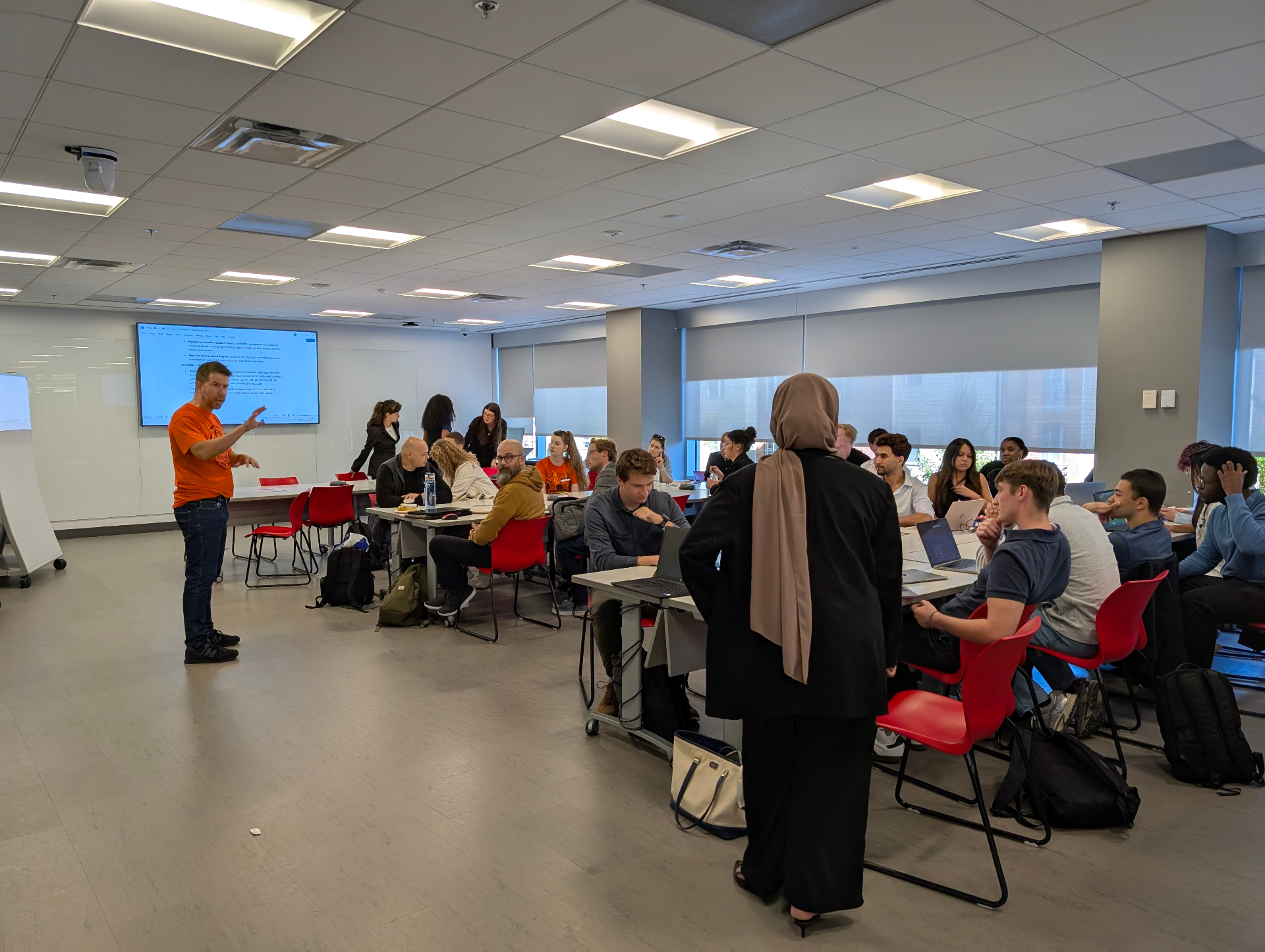 Une salle de classe remplie d’étudiantes, d’étudiants et de membres du corps professoral participe à des discussions en groupe lors d’un atelier. Un animateur en chandail orange s’adresse au groupe à l’avant, tandis que des équipes collaborent autour de tables avec des ordinateurs portables.