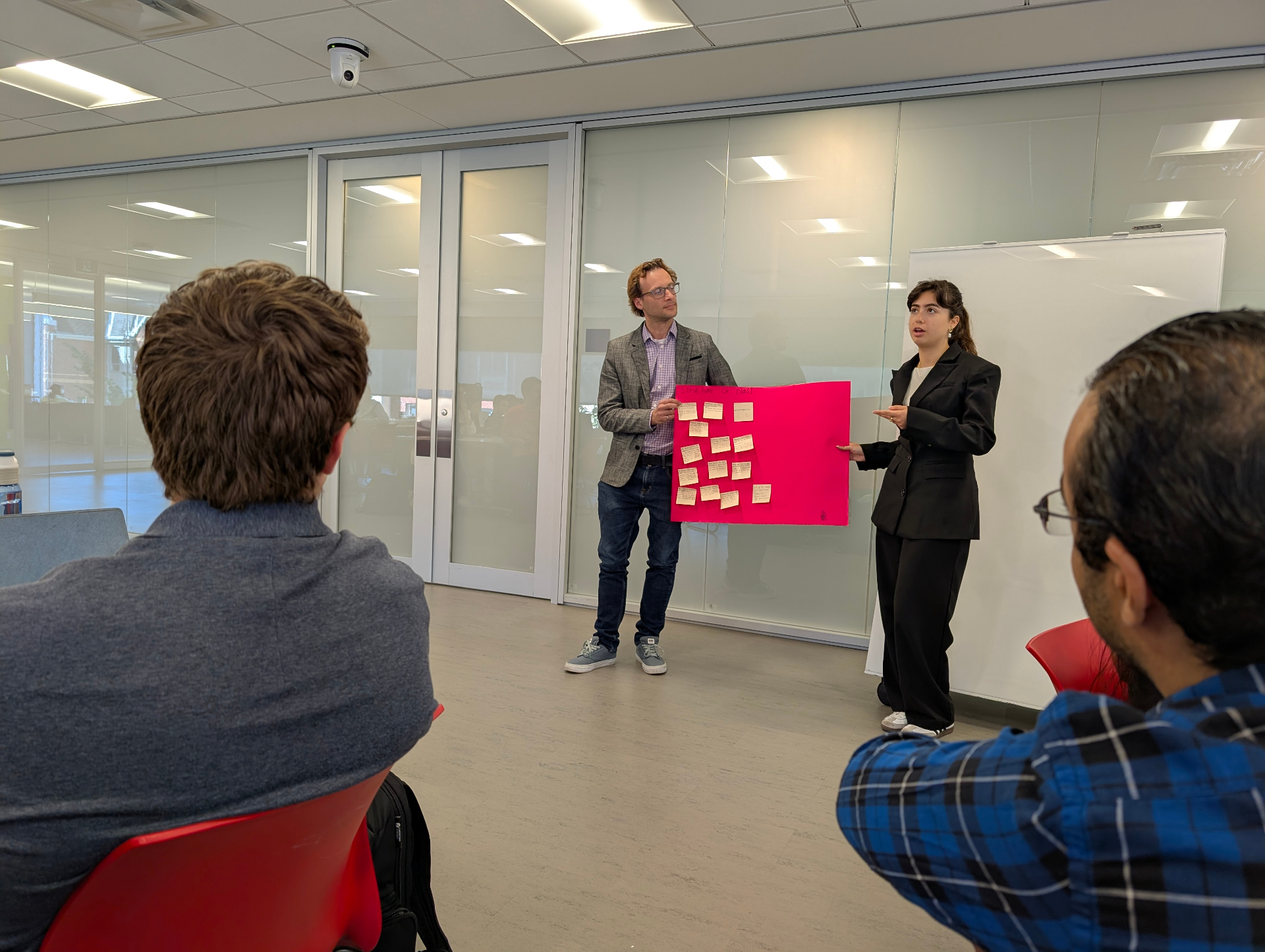 Deux participantes et participants se tiennent à l’avant d’une salle de classe pour présenter les idées de leur groupe à l’aide d’une grande affiche rose recouverte de notes autocollantes, pendant que les autres écoutent attentivement.