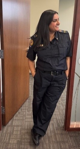 Sunita dressed in a dark official uniform stands casually in a doorway with a smile, hands in her pockets. She appears confident and relaxed in a professional setting.