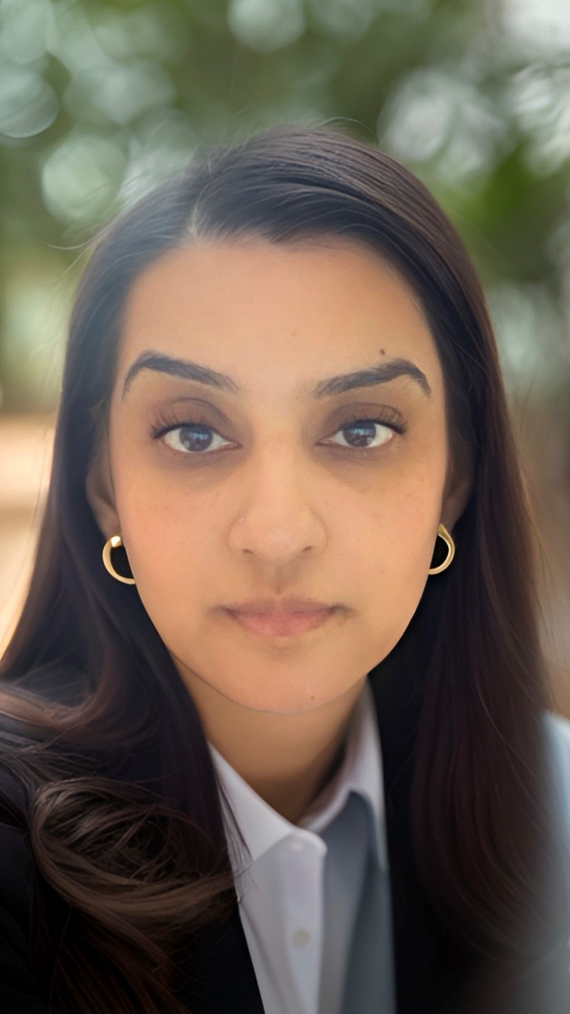 A close-up portrait of Sunita Sekhon, looking directly at the camera with a calm and composed expression. She is wearing a dark blazer and light-colored shirt, with gold hoop earrings. The background is softly blurred with natural green tones.