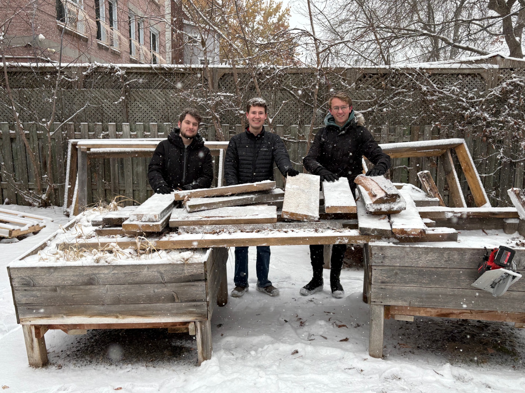 Trois personnes se tiennent dehors dans un jardin communautaire enneigé, souriant en tenant des planches de bois récupérées posées sur des bacs de jardin surélevés. La neige tombe et recouvre le sol et le bois d’une fine couche blanche, avec des arbres et une clôture en arrière-plan.