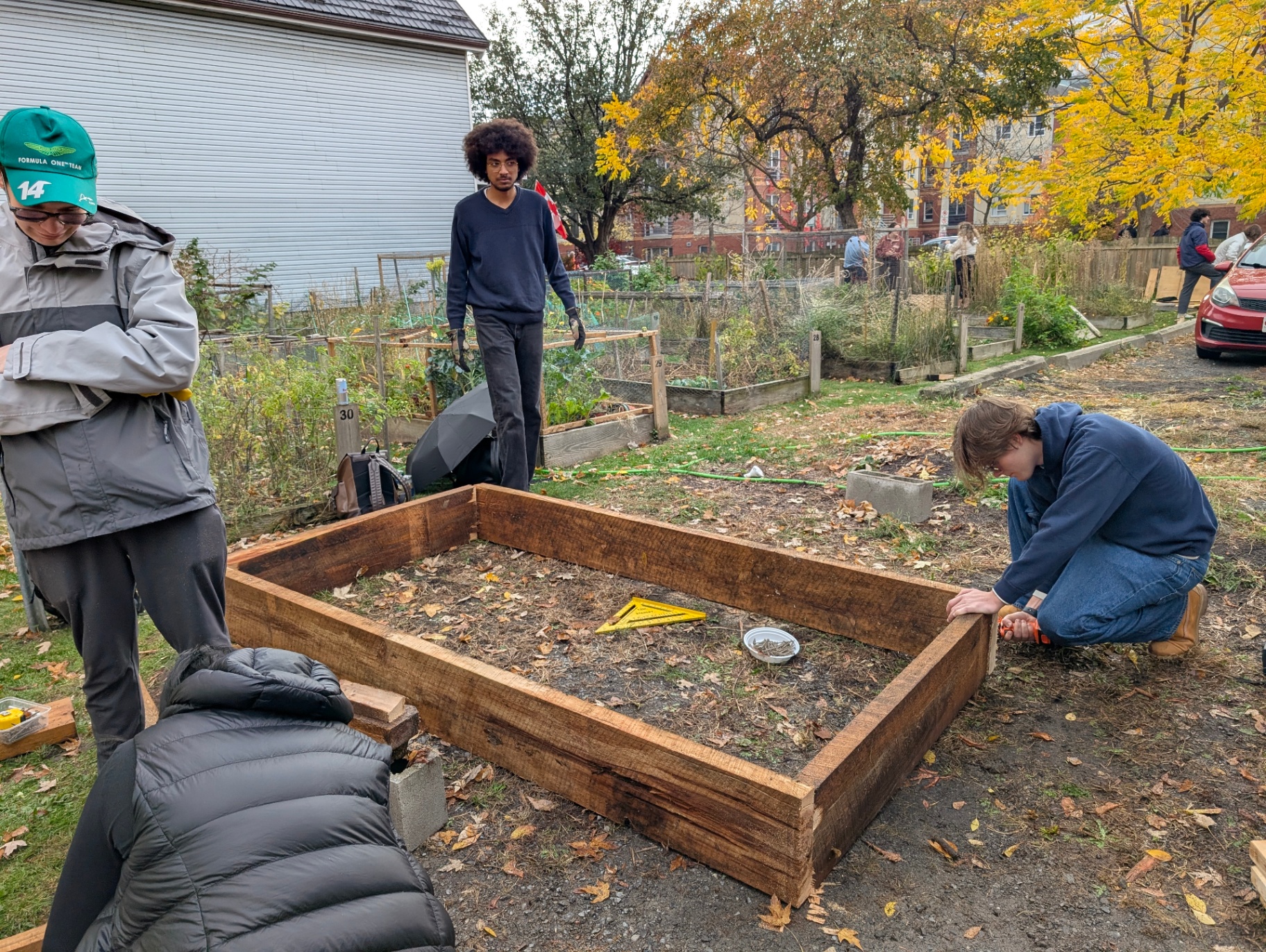 Un groupe de personnes construit un carré de jardin en bois dans un jardin communautaire en automne. Une personne est agenouillée pour aligner un coin du cadre en bois, une autre se tient debout avec des gants, et des feuilles tombées couvrent le sol. Des parcelles de jardin surélevées et des arbres aux feuilles jaunes apparaissent à l’arrière-plan.