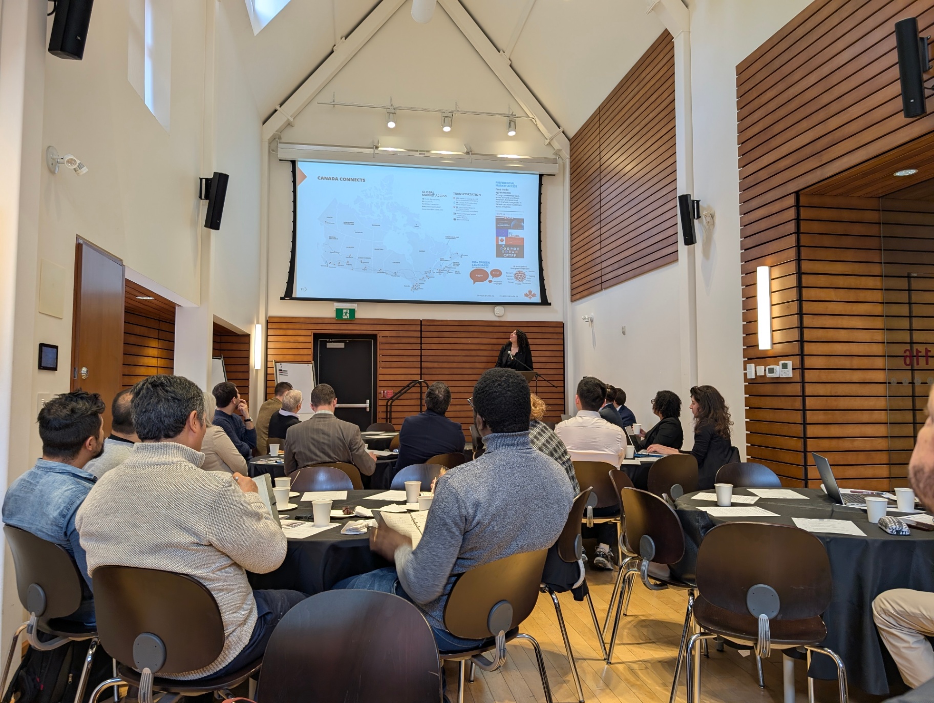 A wide shot of participants seated at round tables in the Alex Trebek belairdirect Hall, listening to a presenter speaking at a podium. A large slide showing a map and trade information is projected at the front of the room.