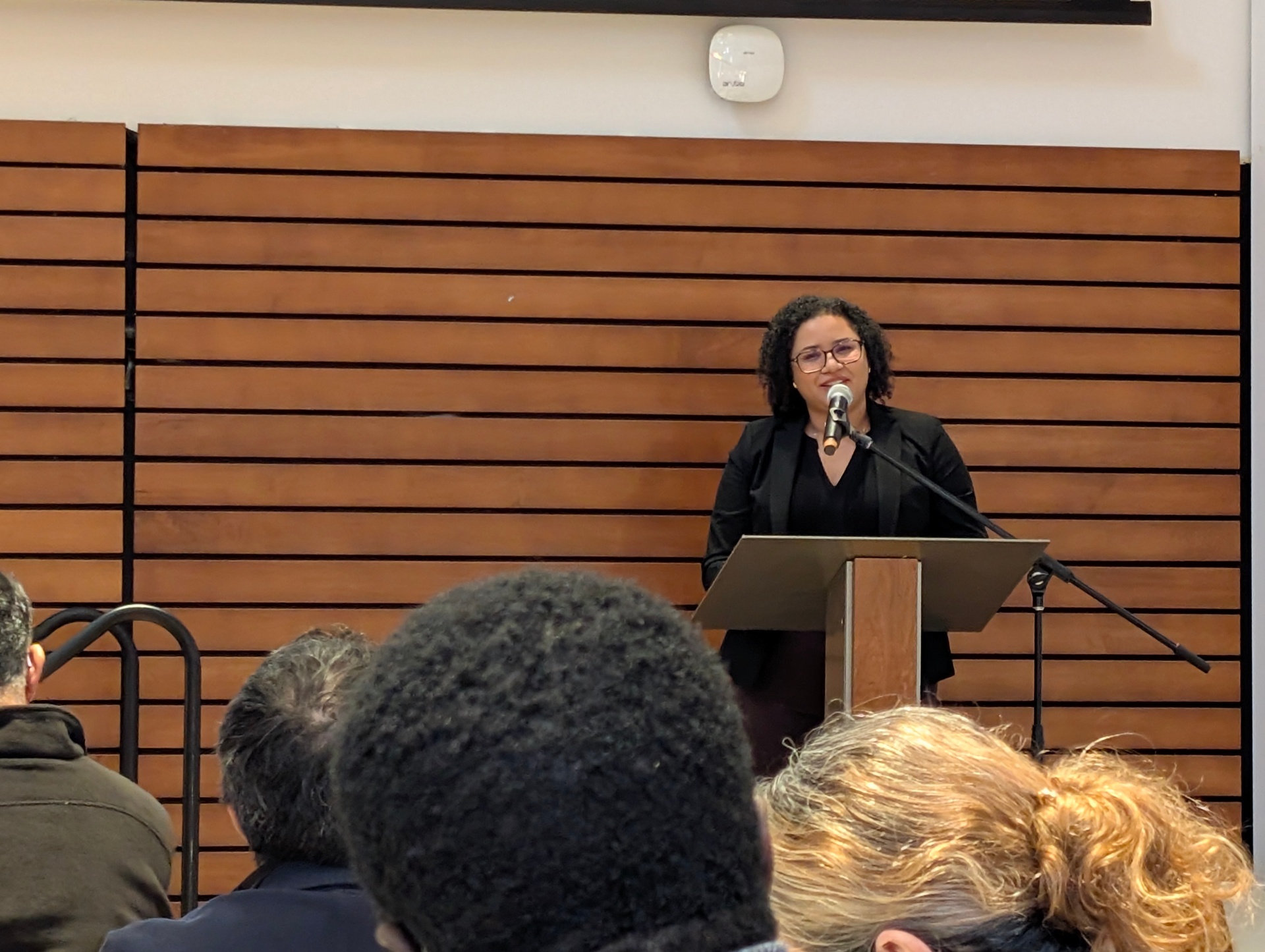 A speaker stands at a podium addressing the audience, with wood-panelled walls and attentive participants visible in the foreground.
