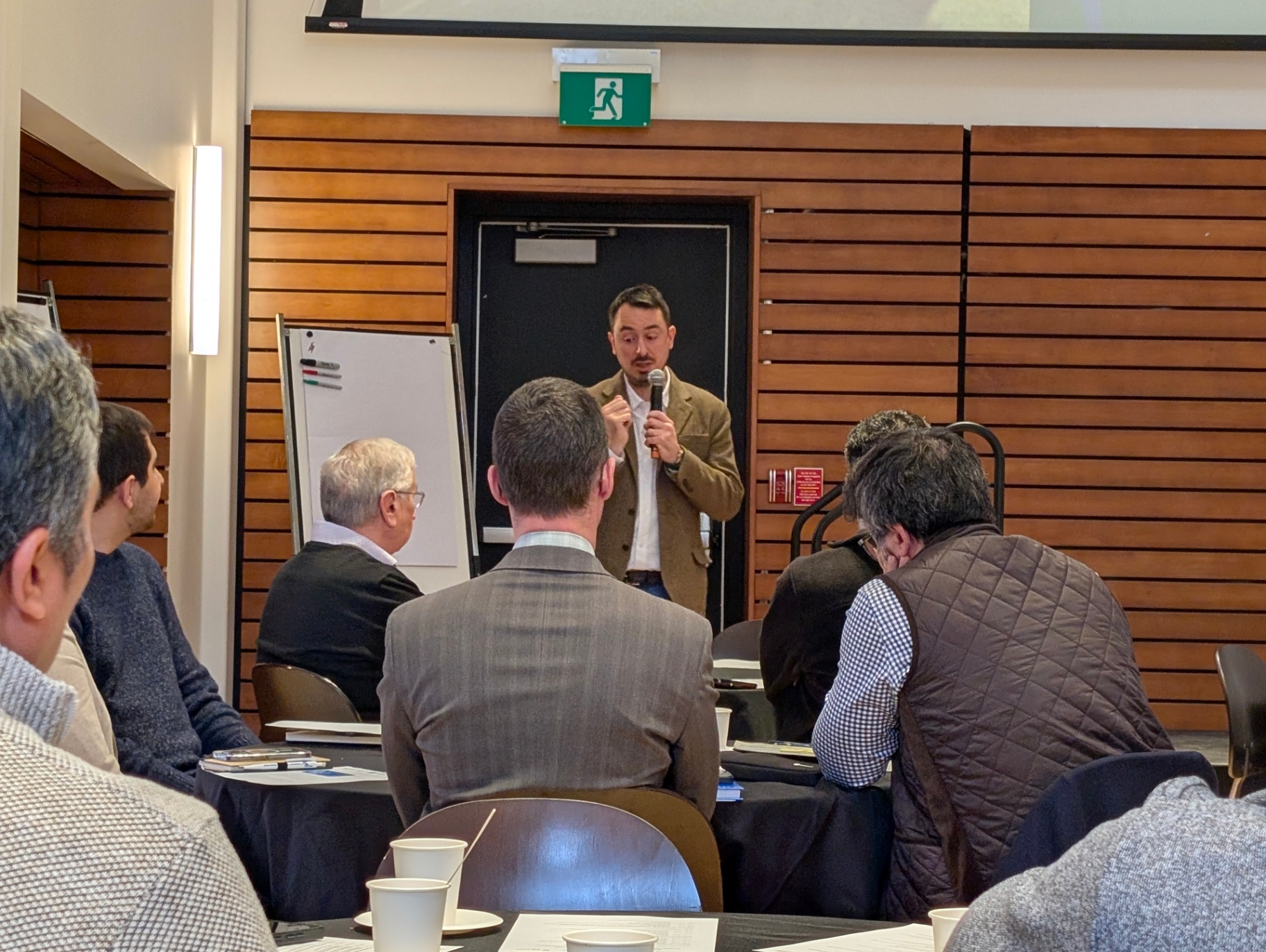 A speaker holding a microphone stands near the back of the room, addressing attendees seated around him. Flip charts and wood-panelled walls are visible in the background.