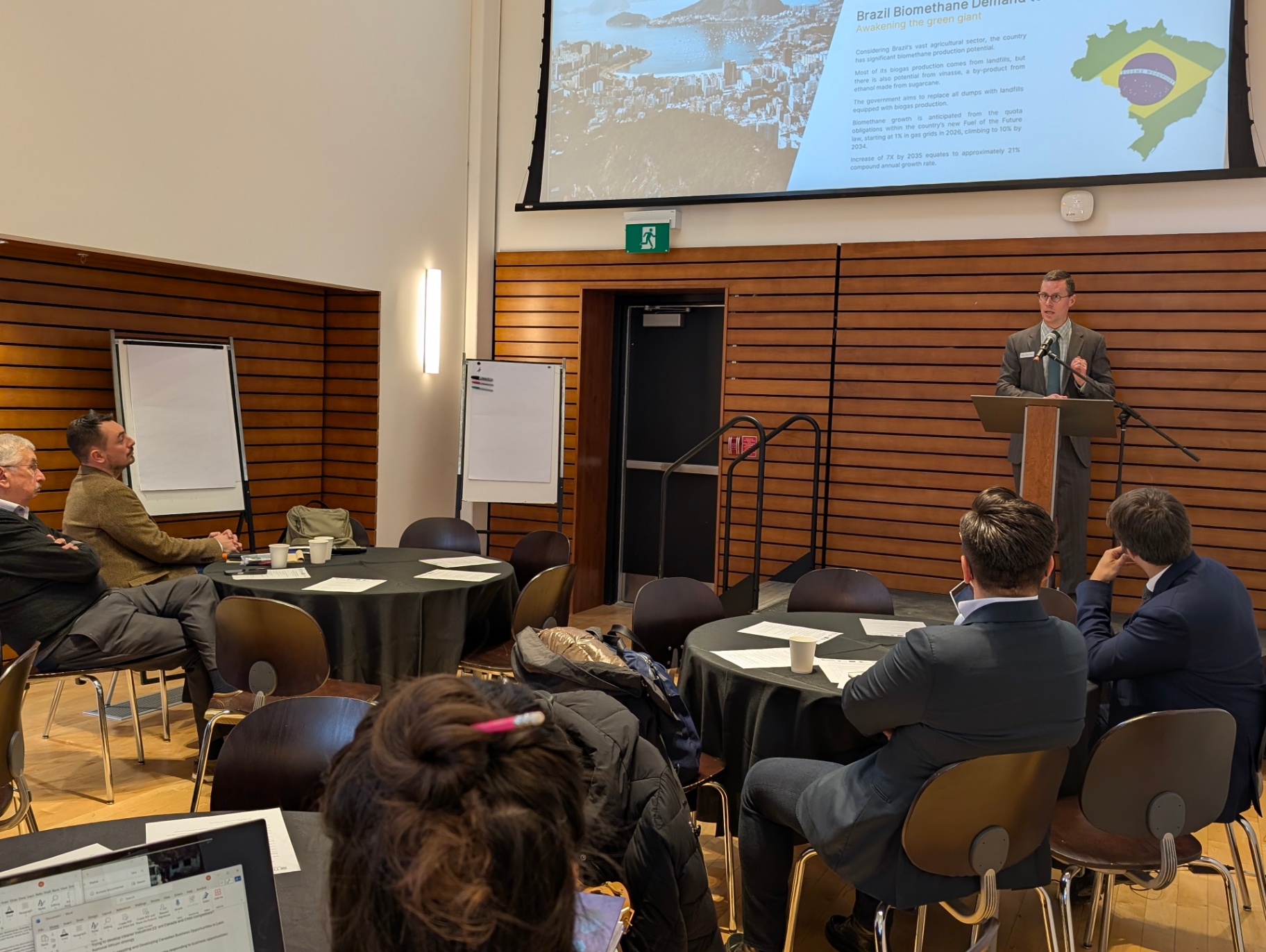 A presenter at the front of the room speaks at a podium while a slide about Brazil’s biomethane sector is projected behind him. Participants sit at round tables facing the stage.