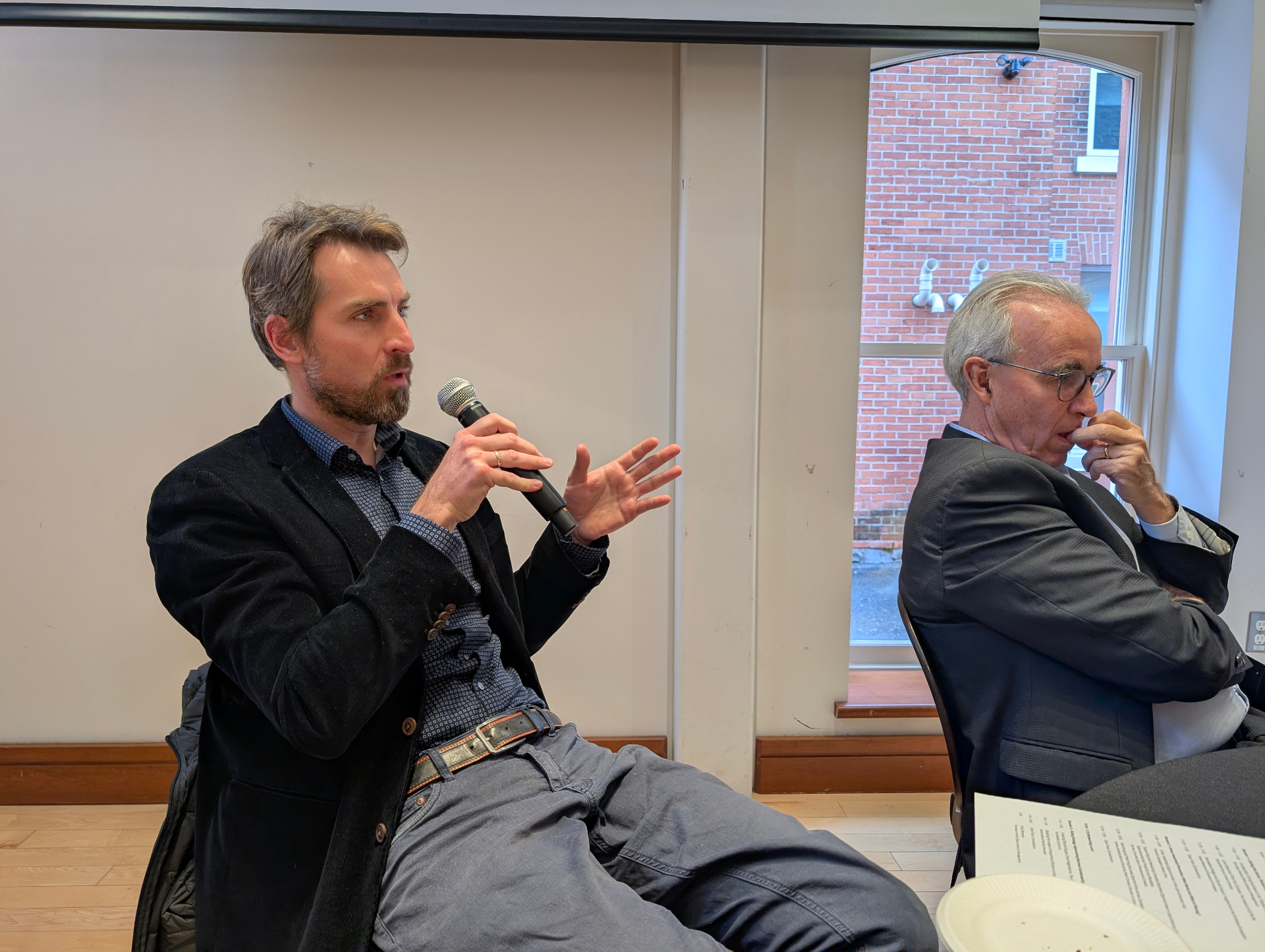 A participant speaks into a microphone while gesturing with his hand during the discussion. Another attendee sits beside him, listening attentively.