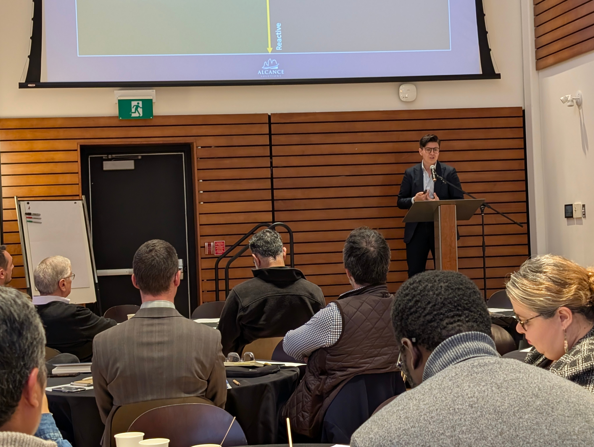 A presenter stands at a podium delivering remarks, with attendees seated and listening. A slide from Alcance Solutions is projected above.