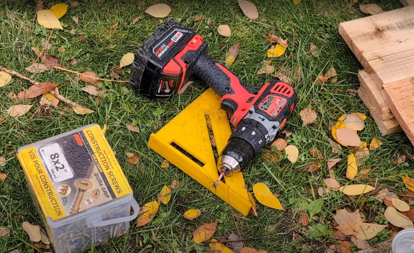 Gros plan de plusieurs outils de construction posés sur de l’herbe couverte de feuilles d’automne, dont une perceuse rouge et noire, une équerre jaune, une boîte de vis de