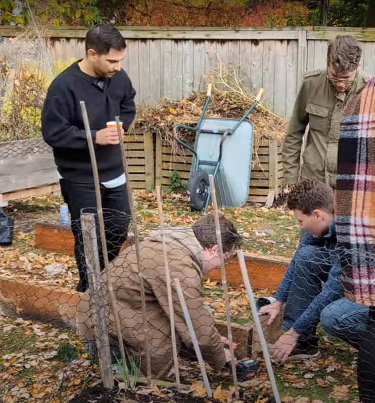 Plusieurs personnes travaillent ensemble dans un jardin communautaire, entourées de feuilles d’automne. Deux individus sont agenouillés pour assembler des morceaux de bois, tandis qu’un autre se tient debout avec une boisson et un quatrième observe. Une brouette et un tas de compost se trouvent près de la clôture en arrière-plan.