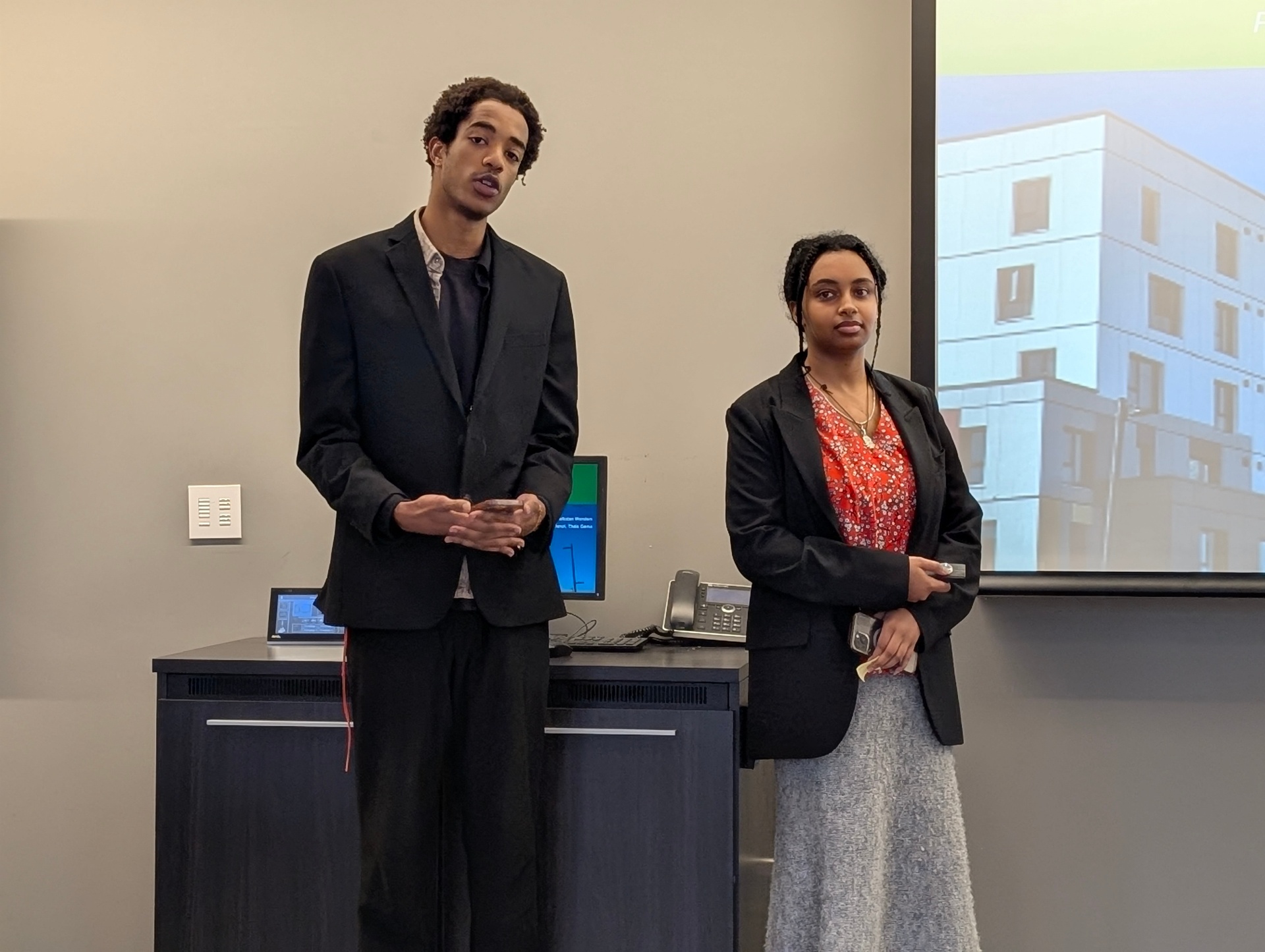 Two students stand side by side presenting their project, speaking to the audience while a building-focused sustainability slide is projected behind them.