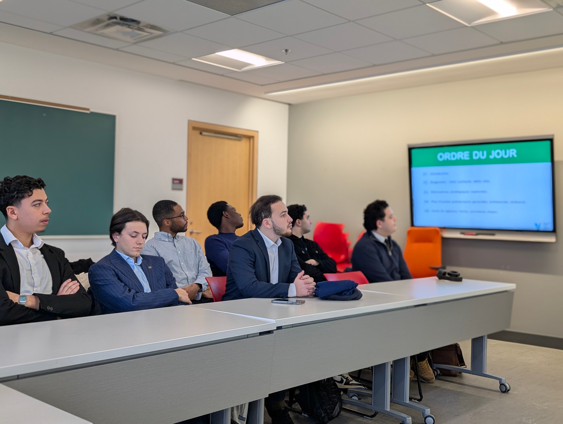 Students seated at tables listen attentively during project presentations in a classroom setting, with a screen displaying the agenda in the background.
