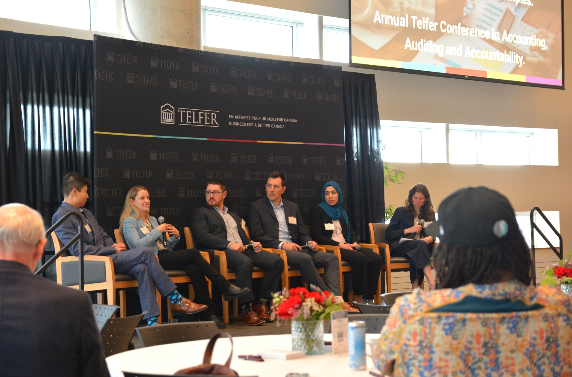 Six panelists, three men and three women, sit in chairs on stage as they address the audience.