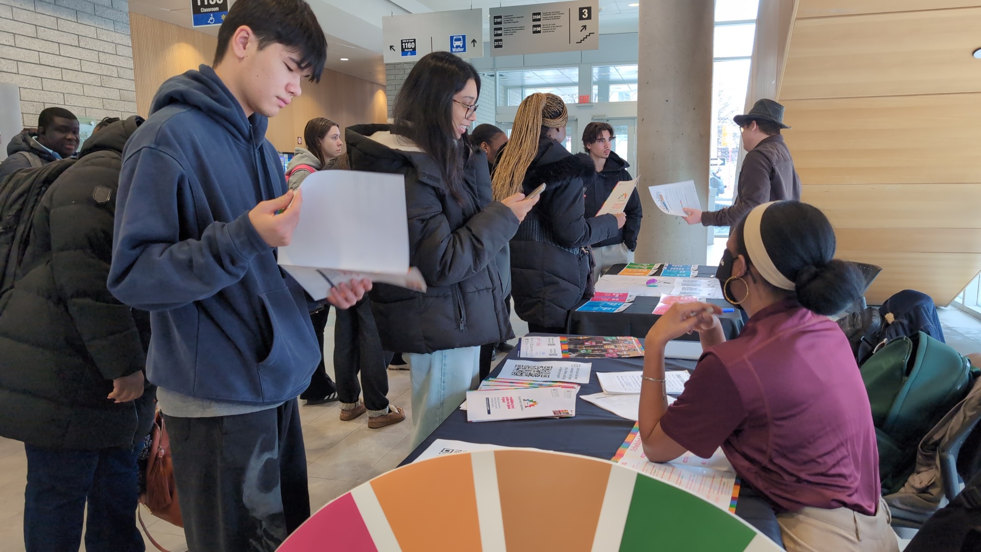 Students interact at an information table on campus, reviewing materials and asking questions while a staff member provides guidance, with SDG-themed resources visible.