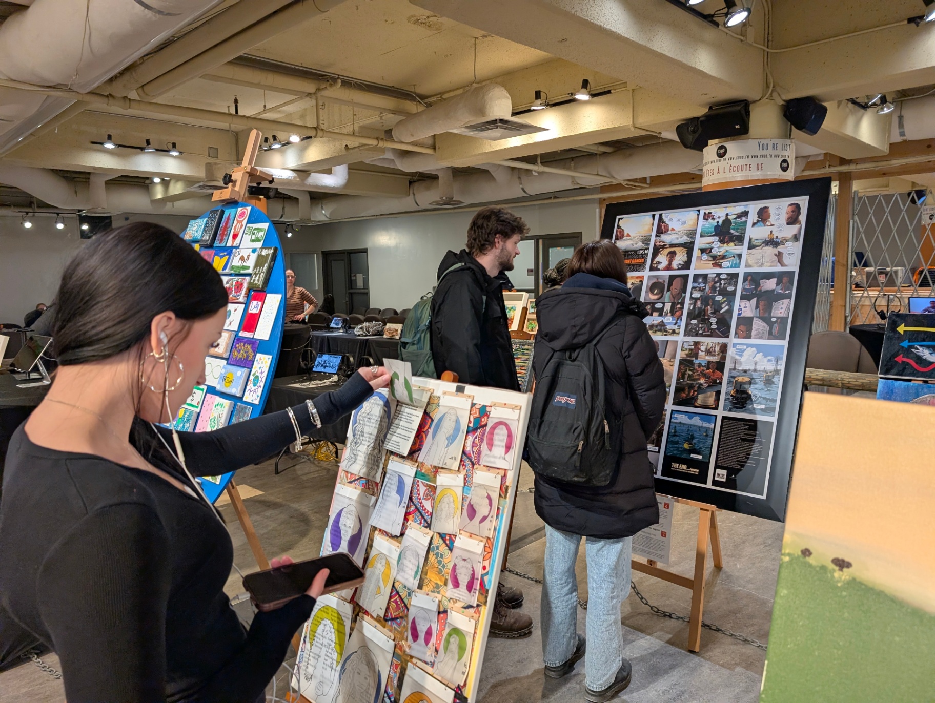 Students explore a campus art exhibition, viewing illustrated portraits and photo displays mounted on boards, while engaging with creative works in an indoor gallery setting.