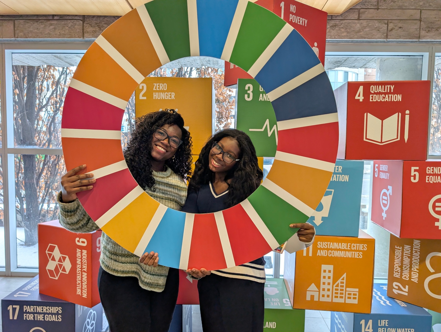 Two students smile while holding a large circular Sustainable Development Goals (SDG) wheel, standing in front of colorful SDG display cubes highlighting goals such as quality education, gender equality, and sustainable cities.