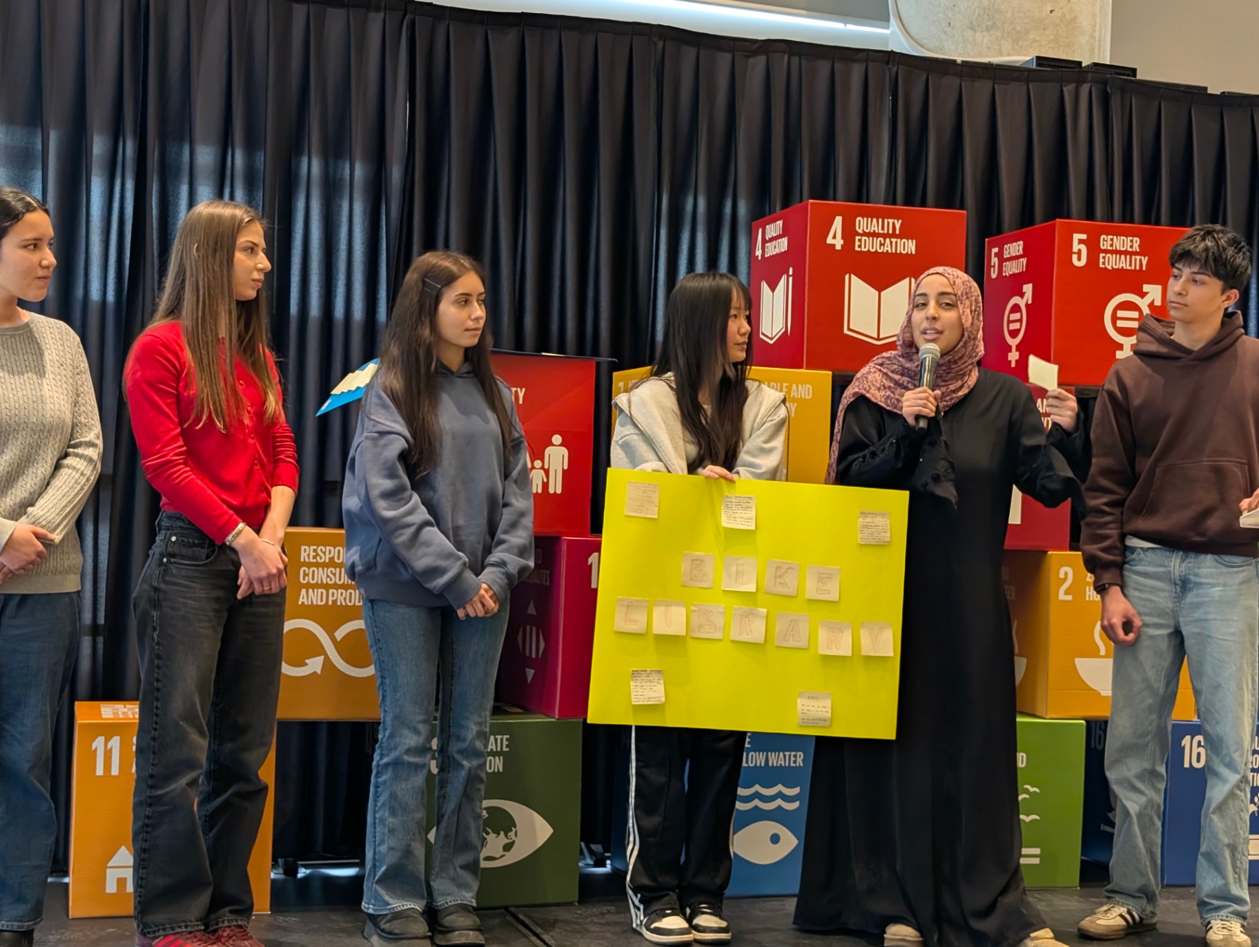 A group of students presents on stage, with one student speaking into a microphone while others stand beside a poster board, in front of large SDG cubes highlighting goals such as quality education and gender equality.