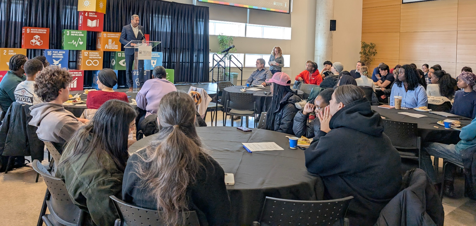 Students sit at round tables listening to a speaker at a podium during a campus event, with large Sustainable Development Goals (SDG) cubes displayed behind the stage.