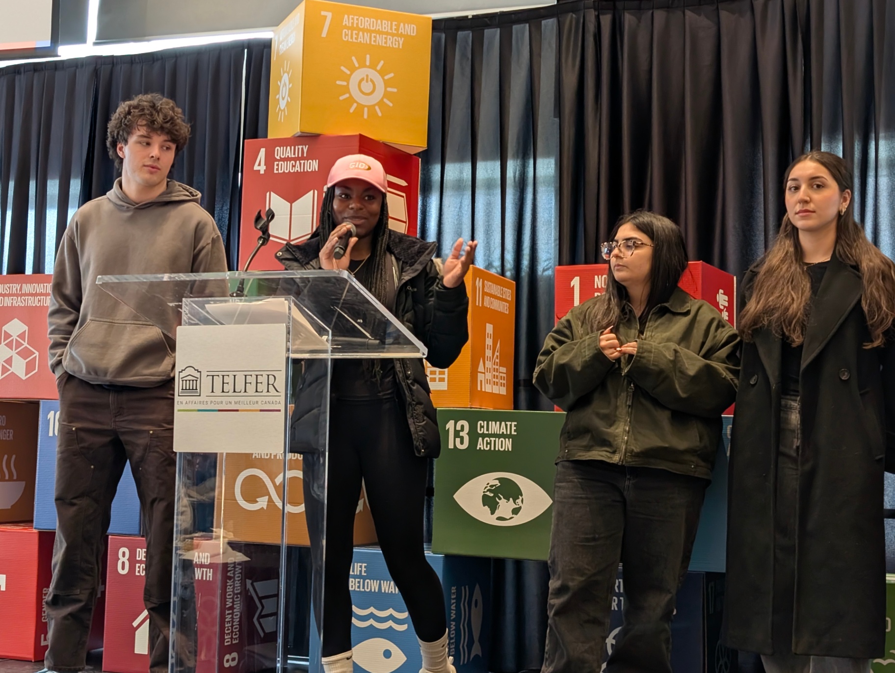 A student speaks into a microphone at a podium during a campus event, while teammates stand beside her in front of large Sustainable Development Goals (SDG) cubes.
