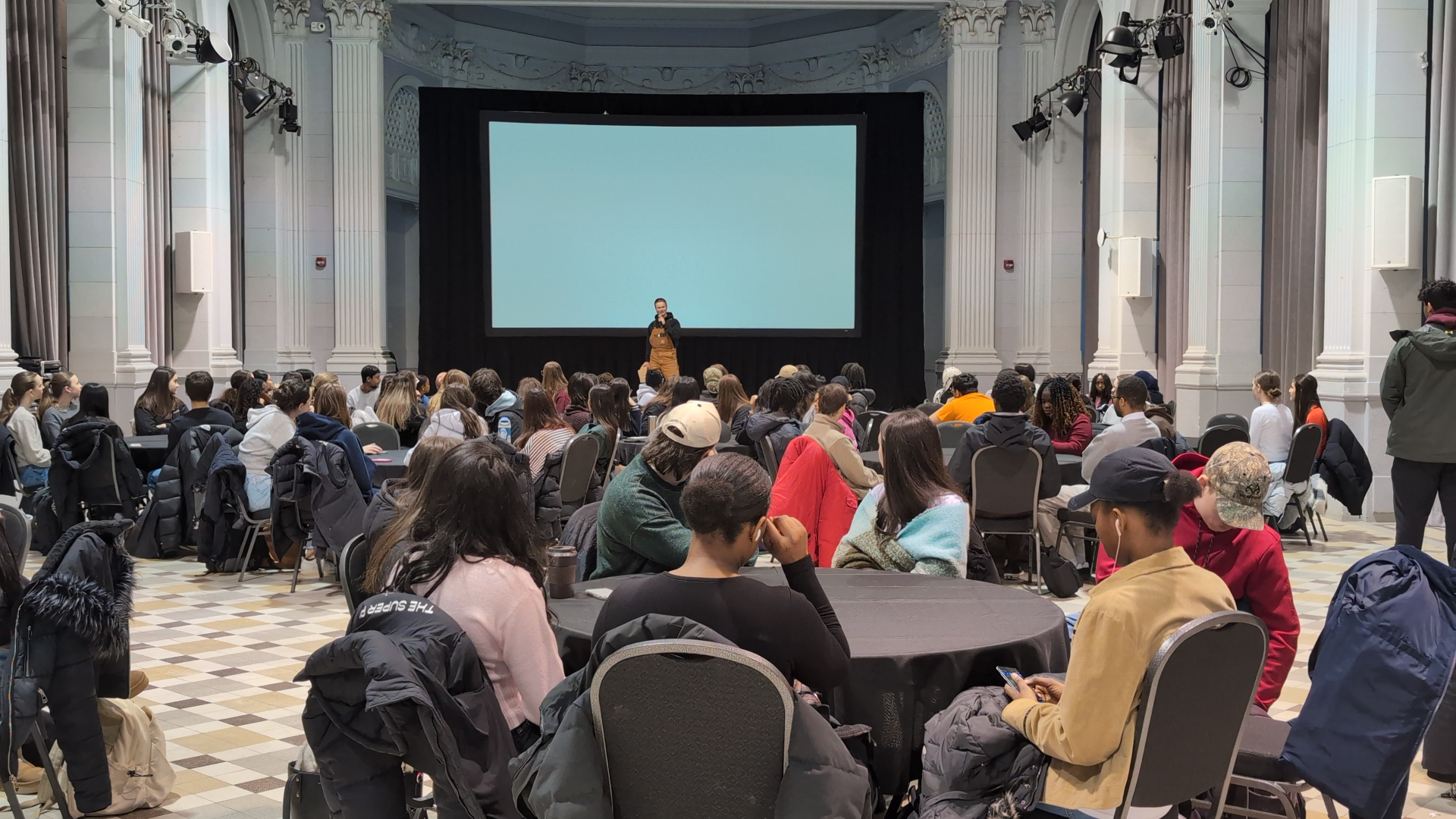A large audience of students sits at round tables in a hall, listening to a speaker on stage in front of a large screen during a campus event.