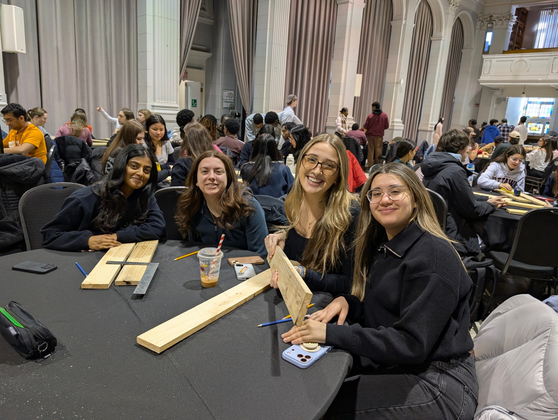 A group of students sits around a table smiling and working with wooden materials during a hands-on workshop, with tools and pieces of wood laid out in front of them.