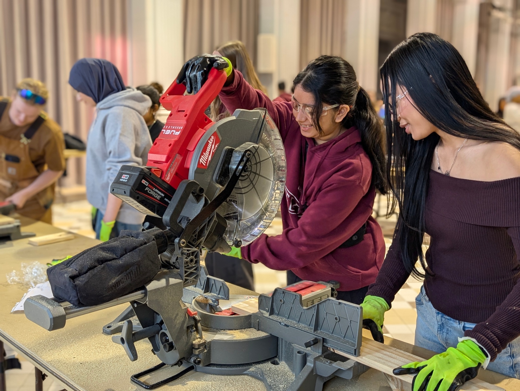 Two students wearing safety glasses and gloves use a power saw to cut wood during a hands-on workshop, while others work in the background.