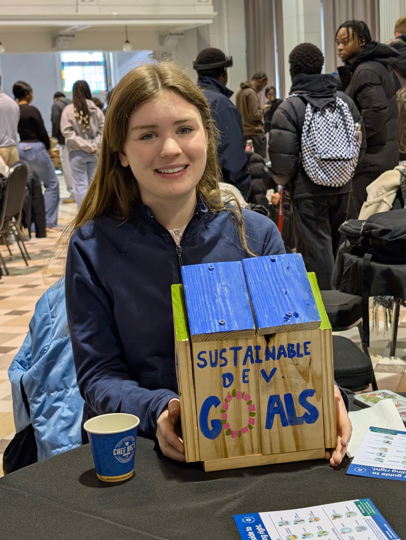 A student smiles while holding a small handmade wooden birdhouse painted with the words &ldquo;Sustainable Dev Goals&rdquo; during a workshop, with other participants in the background.