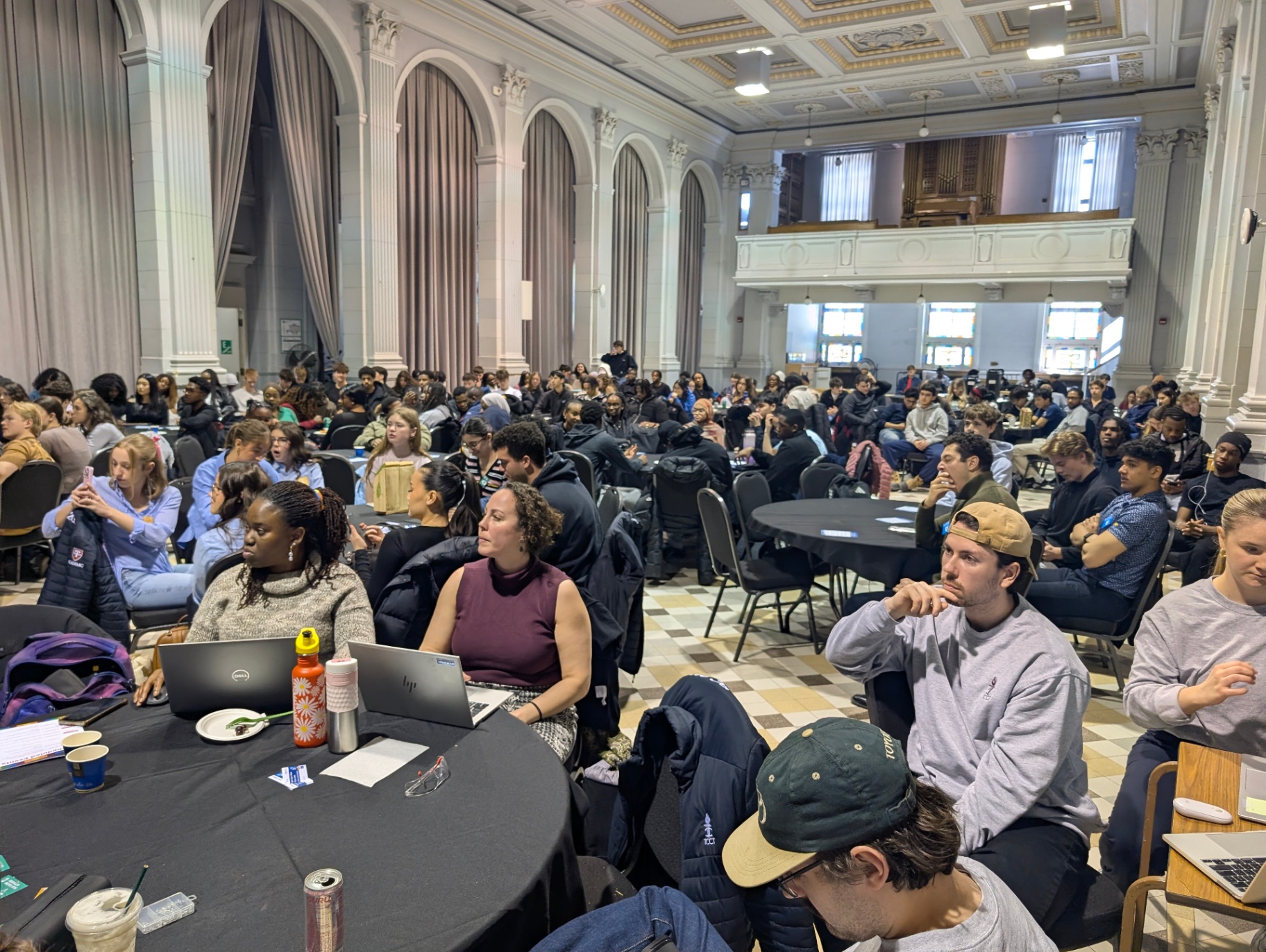 A large group of students fills a grand hall, seated at round tables and engaged in a campus event, some listening attentively while others take notes or use laptops.