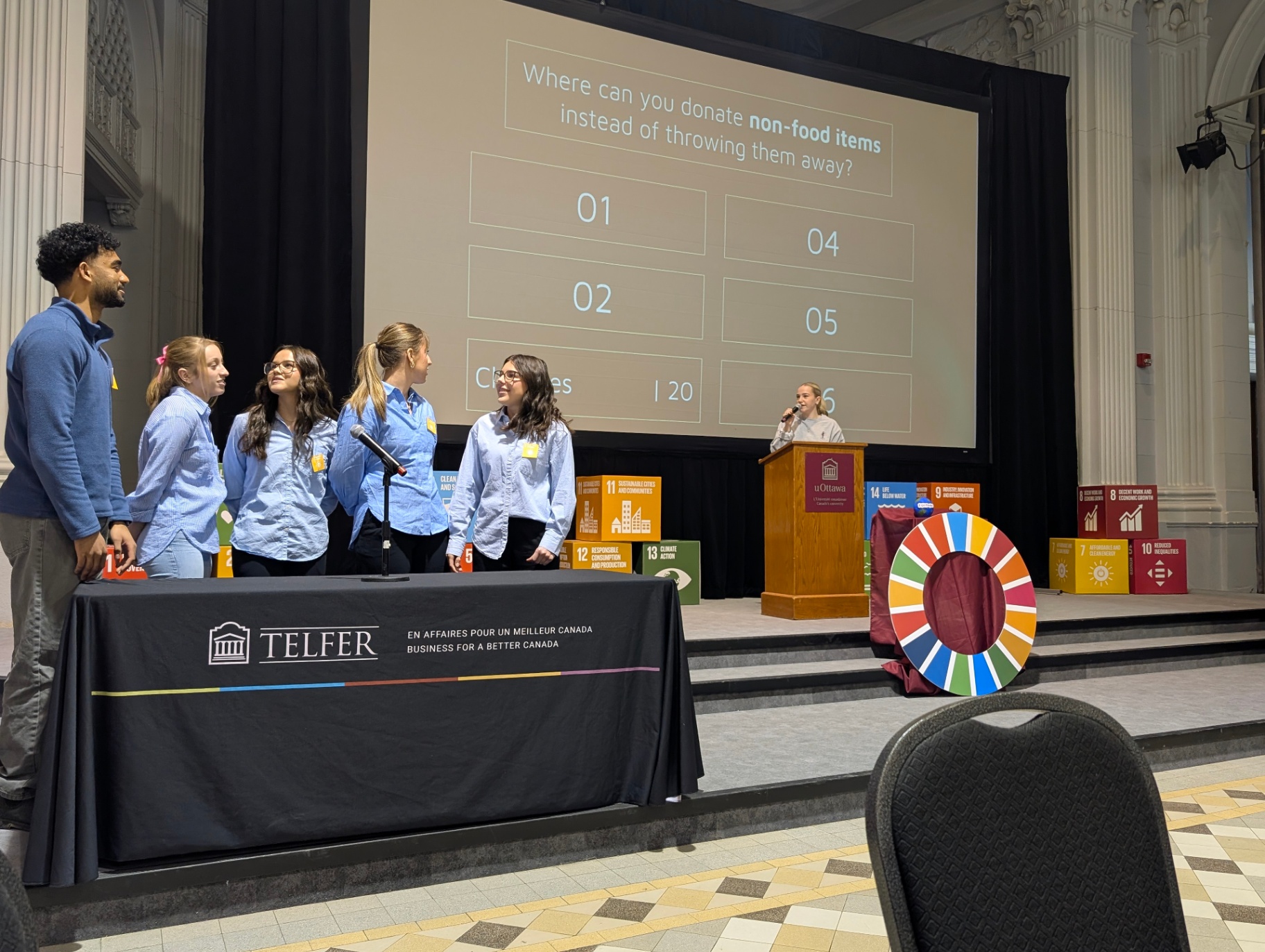A group of students stands on stage during a sustainability-themed event, participating in an interactive quiz while a presenter speaks at a podium and a large screen displays a question about donating non-food items.
