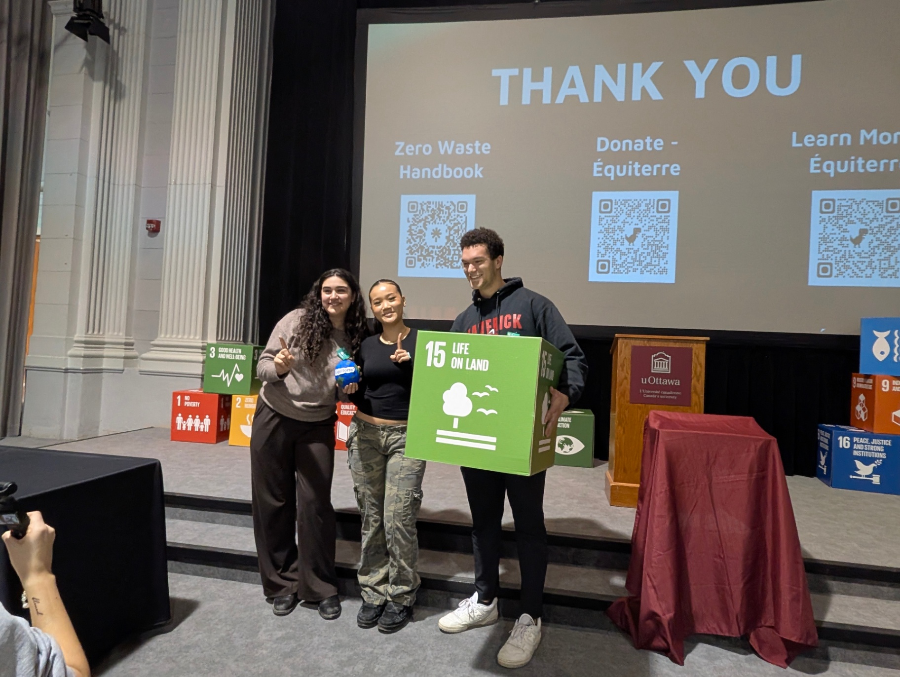 Three students pose on stage holding a large SDG 15 &ldquo;Life on Land&rdquo; cube after a sustainability event, with a screen behind them displaying &ldquo;Thank You&rdquo; and QR codes.