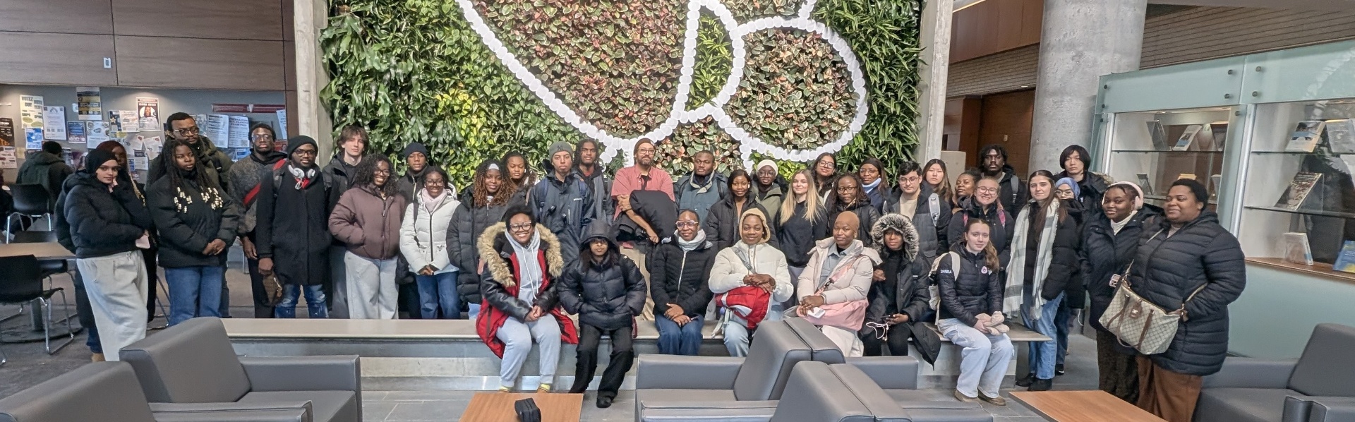 A large group of students poses together in a campus atrium in front of a tall living green wall shaped with a white floral design.