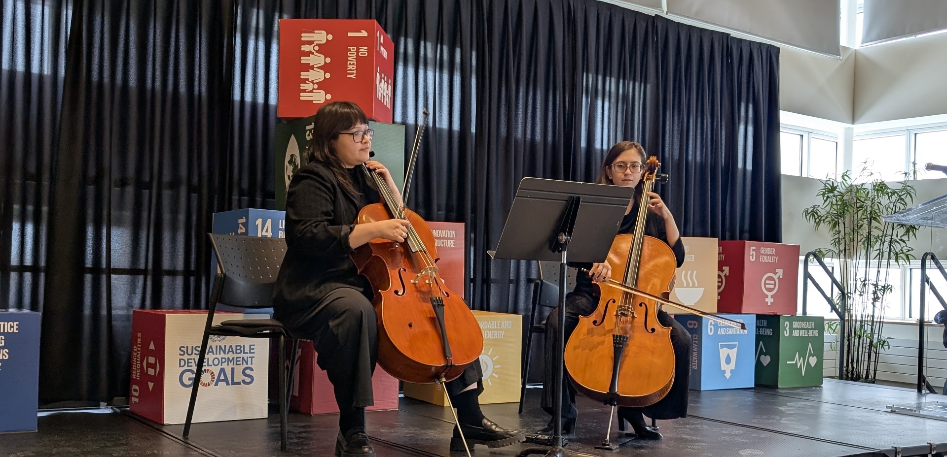 Two musicians perform on cello on stage during a sustainability event, with Sustainable Development Goals cubes displayed behind them.