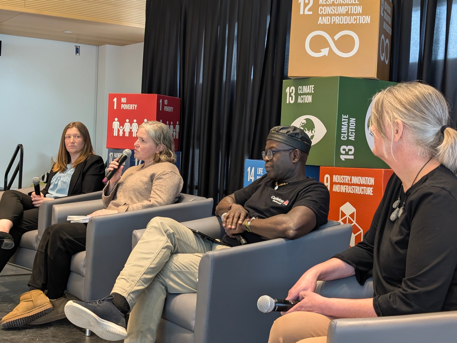 A panel of speakers sits on stage during a sustainability discussion, with Sustainable Development Goals cubes displayed behind them as one speaker addresses the audience.