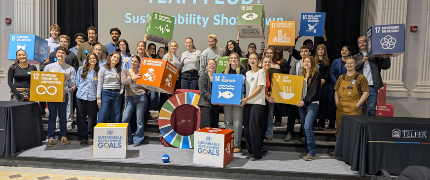 Un grand groupe d’étudiants se tient sur scène en tenant des cubes colorés des Objectifs de développement durable lors d’un événement « Team Feud », posant ensemble devant un écran.