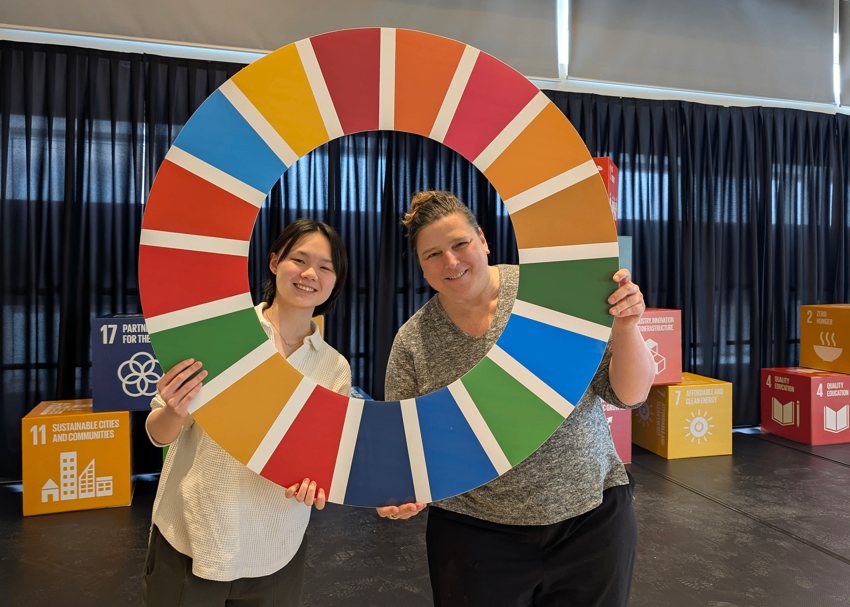 Two participants smile while holding a large circular Sustainable Development Goals color wheel on stage, with SDG cubes displayed in the background.