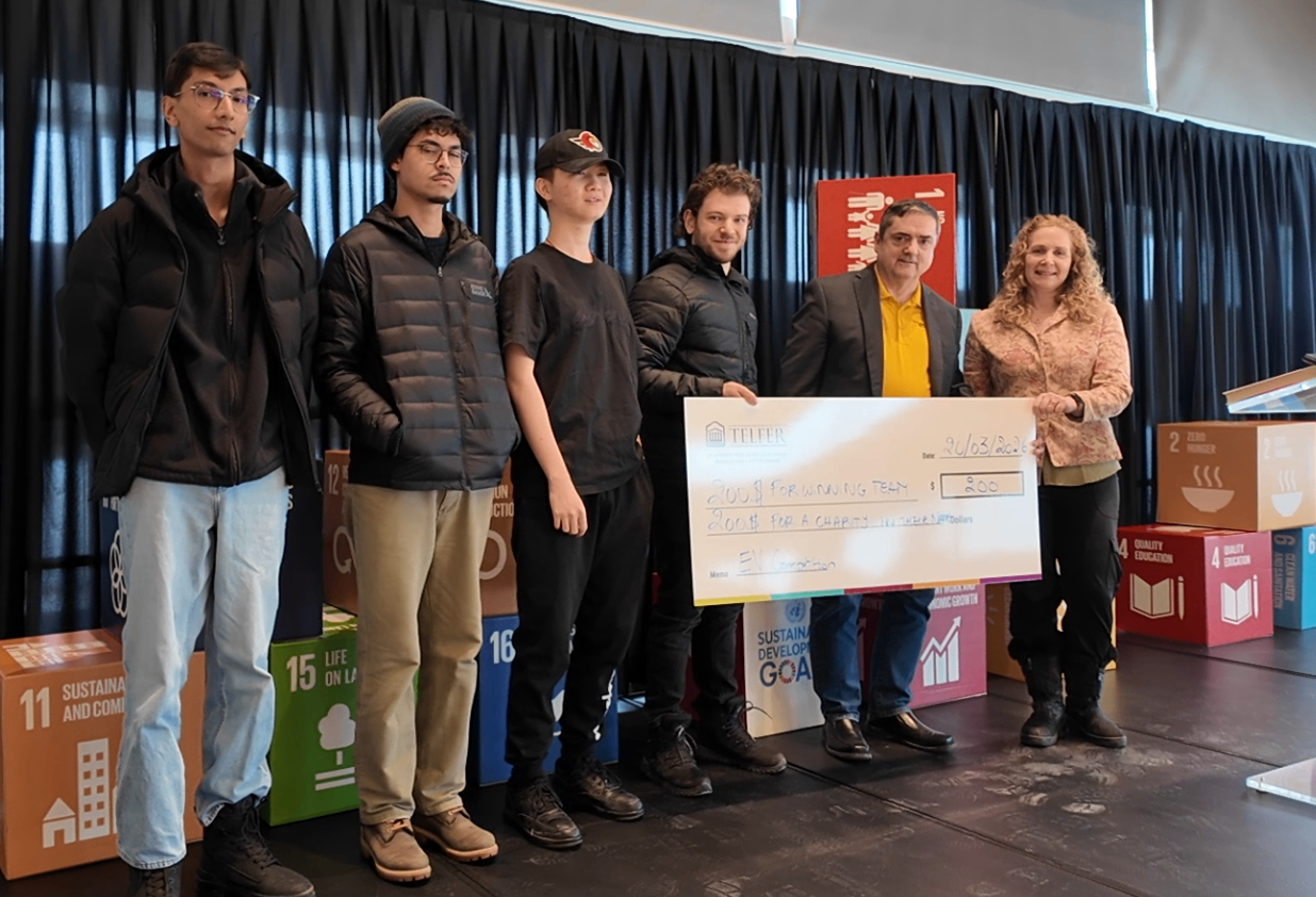 A group of students stands on stage alongside two organizers, holding a large cheque awarded to a winning team during a sustainability event, with SDG cubes displayed behind them.