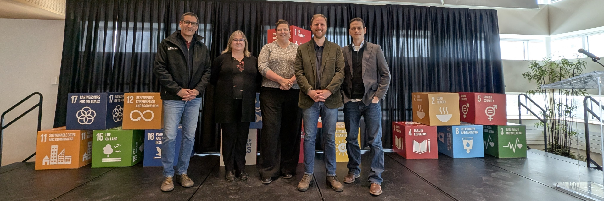 A group of speakers and organizers stands together on stage during a sustainability event, with Sustainable Development Goals cubes displayed behind them.