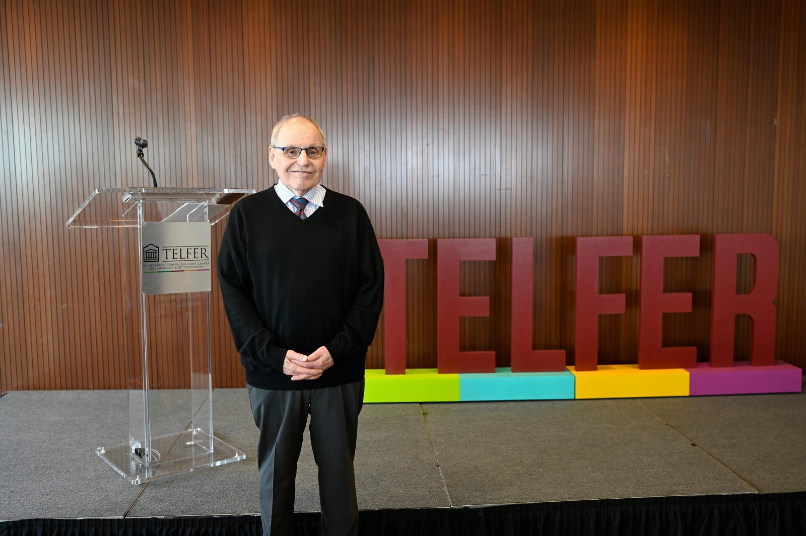 Portrait of Julian Barling standing on stage beside a podium at the Telfer School of Management, with large Telfer letters displayed in the background