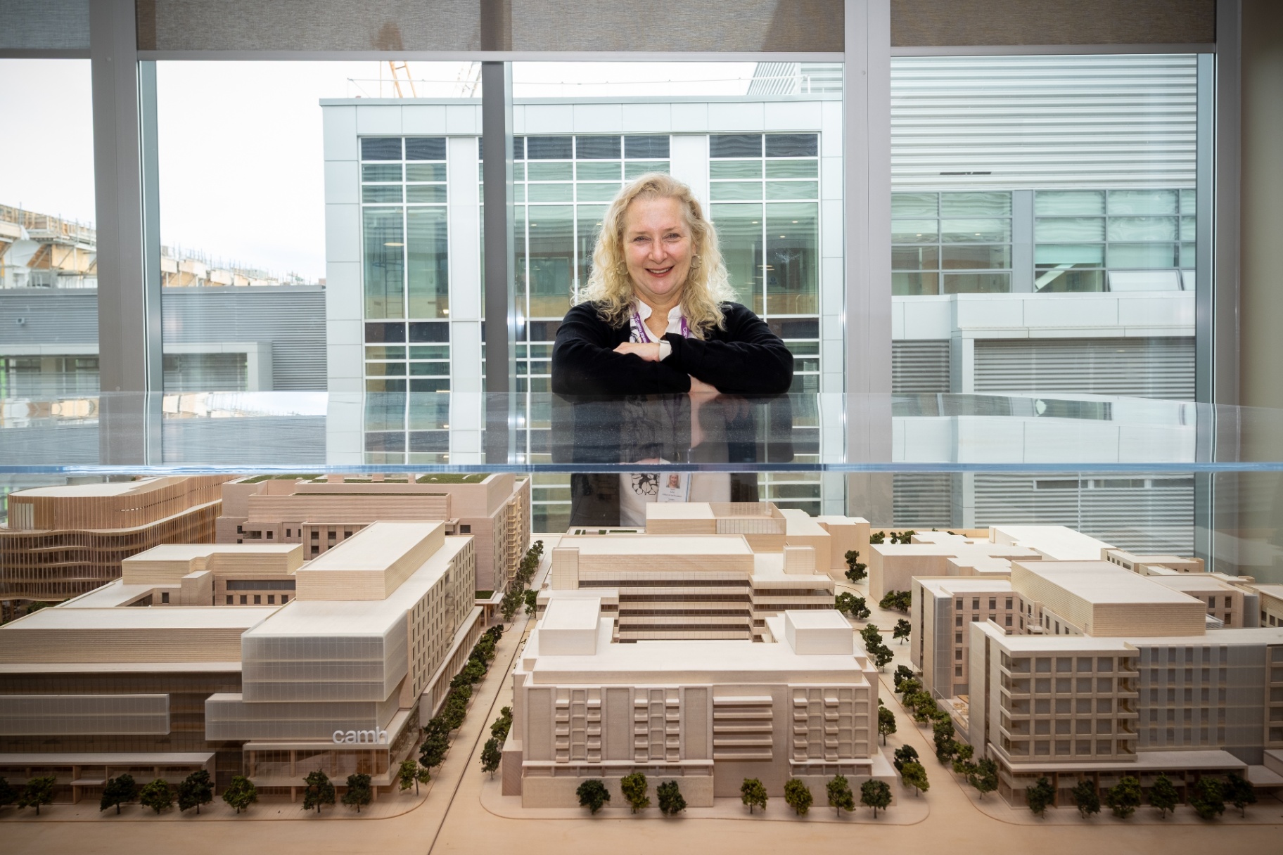 Sarah Downey smiles and leans forward with arms crossed over a detailed architectural scale model of the CAMH campus, with large windows in the background.